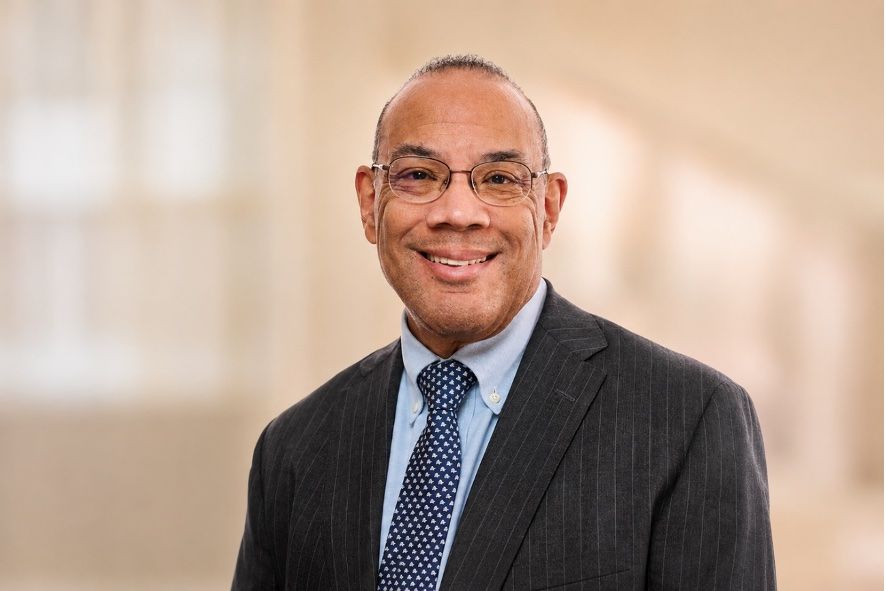 A man in a pinstripe suit, blue shirt, and dark blue tie smiles for a portrait in front of a neutral background.