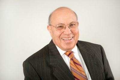 A man in a dark suit, white shirt and striped orange tie smiles for a portrait in front of a neutral background.