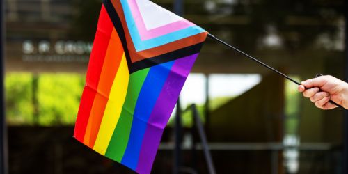 A light skinned hand holds a pride flag that includes rainbow stripes and chevrons. This flag is held in front of a slightly blurred Crown Hall building on Illinois Tech campus in the background 