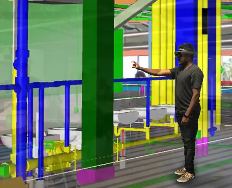 Rendering of virtual reality building with systems such as water pipes highlighted in different colors. A person is depicted reaching out in front of them to interact with the simulation.